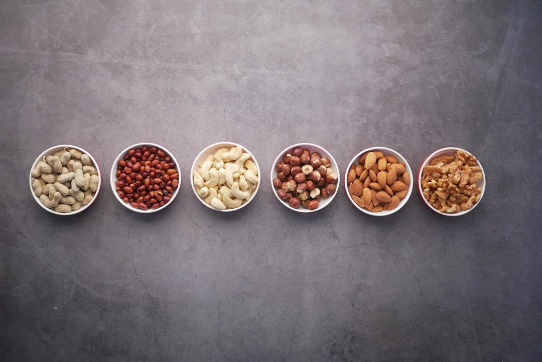A rustic wooden board displaying artisan roasted almonds, pecan halves, macadamia nuts, and a jar of craft nut butter beside an open gift tin, warm natural light